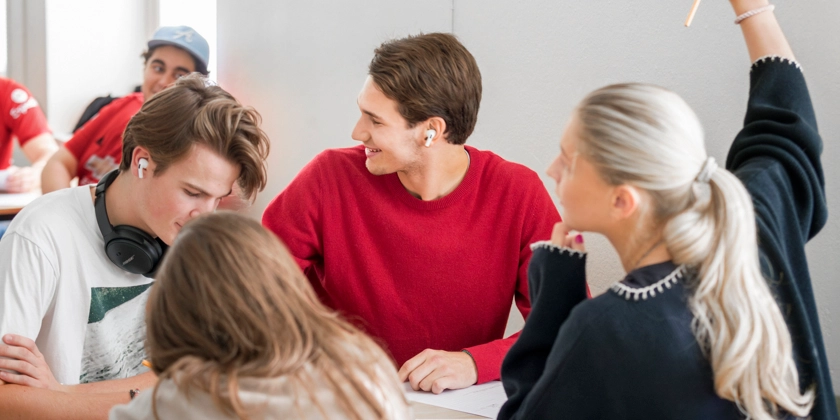 En grupp elever vid ett bord, en av dem räcker upp handen. 