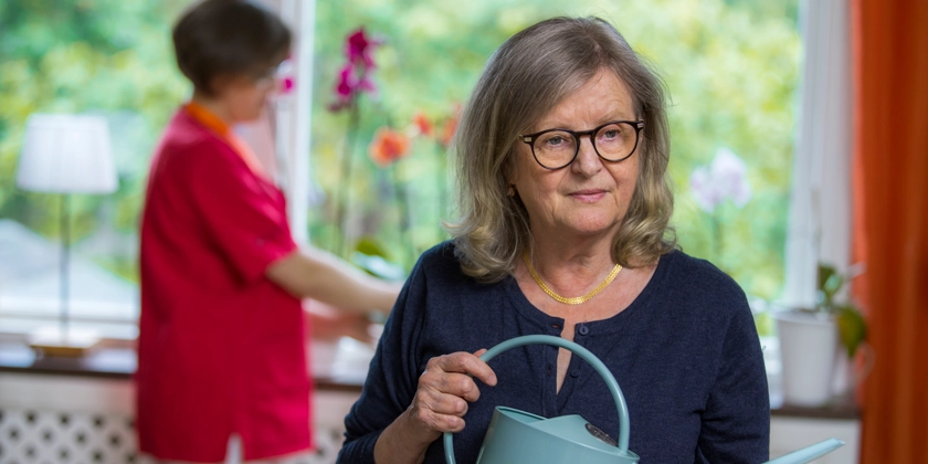 En äldre kvinna håller en vattenkanna medan en annan kvinna pysslar med blommor i fönstret.