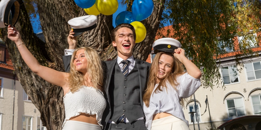 En grupp glada studenter står bredvid varandra framför ett träd vid en skolbyggnad.