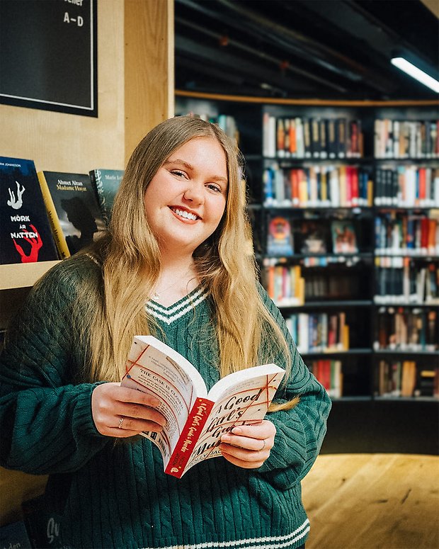 Anna Tärnvik står lutad mot en bokhylla på Lidingö stadsbibliotek. Hon håller ett exemplar av boken »A Good Girl's Guide To Murder« av Holly Jackson.