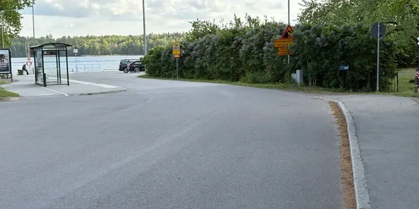 Asfalterad gata med växtlighet till höger och en busskur till vänster i bild och en kaj och vattnet rakt fram .