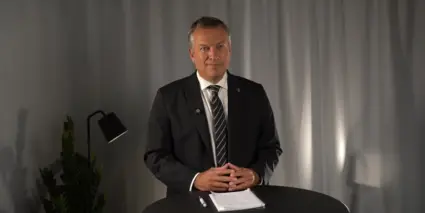 Daniel Källenfors står i kostym vid ett bord med ett blocka och en penna framför sig. Han håller händerna knäppta framför sig och tittar in i kameran