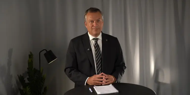 Daniel Källenfors står i kostym vid ett bord med ett blocka och en penna framför sig. Han håller händerna knäppta framför sig och tittar in i kameran