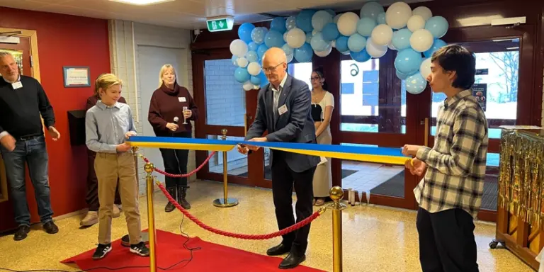 Man klipper band på Lillåkers skola med personer som tittar på. I bakgrunden sitter ballonger och på golvet ligger en röd matta