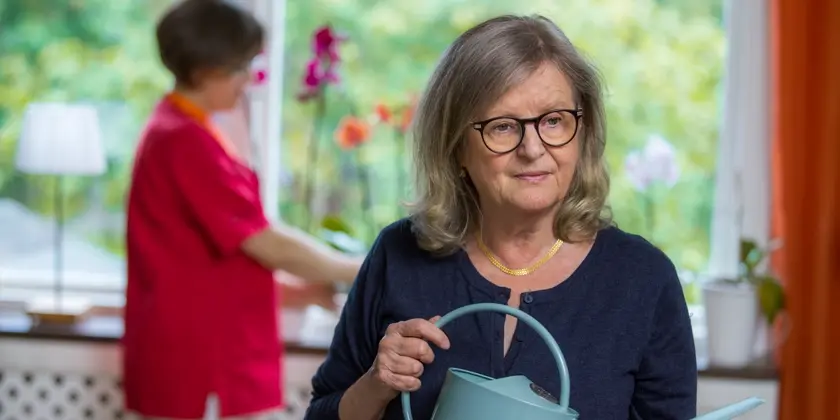 En äldre kvinna håller en vattenkanna medan en annan kvinna pysslar med blommor i fönstret.
