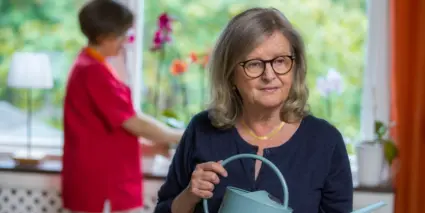 En äldre kvinna håller en vattenkanna medan en annan kvinna pysslar med blommor i fönstret.