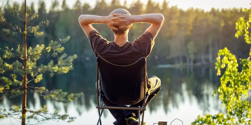 Man sitter vid en sjö i en campingstol med händerna knäppta bakom huvudet och tittar ut över en sjö