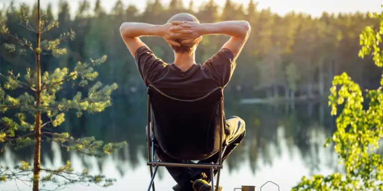 Man sitter vid en sjö i en campingstol med händerna knäppta bakom huvudet och tittar ut över en sjö