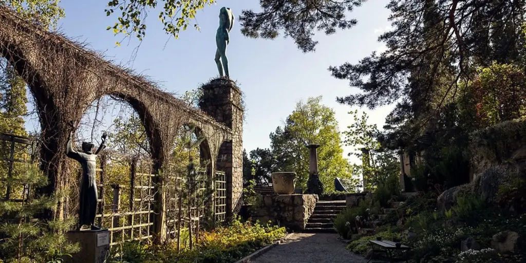 Exteriör från Millesgården. En mur med valv till vänster och en hög staty på en pelare. Fullt med grönska runt om och en grusgång. 