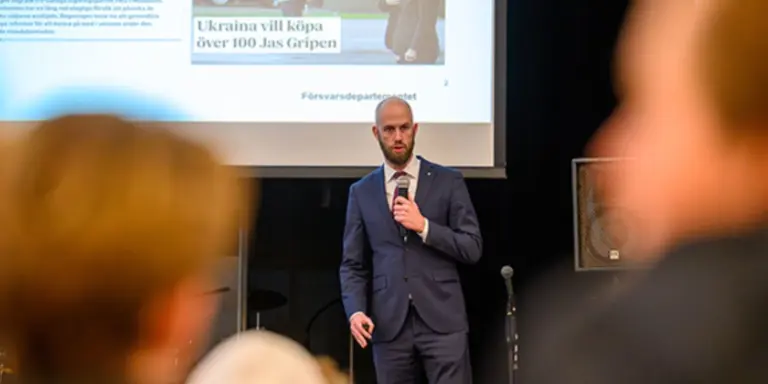 Carl-Oskar Bohlin (M), minister för civilt försvar, står i kostym på scenen och pratar i en mikrofon. I förgrunden synd suddiga åhörare