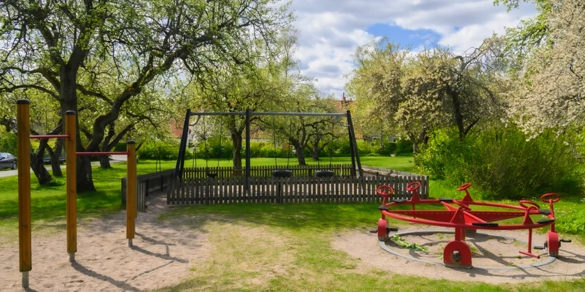 En lekpark med gungställning och cykelsnurra omgiven av grönska.