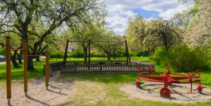 En lekpark med gungställning och cykelsnurra omgiven av grönska.