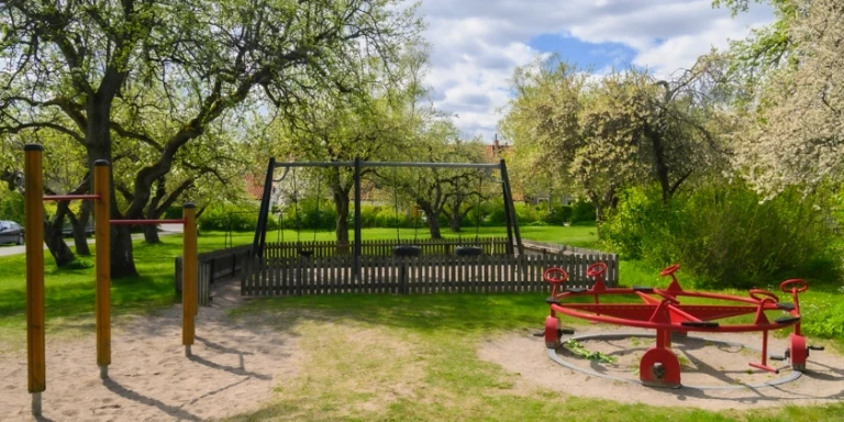 En lekpark med gungställning och cykelsnurra omgiven av grönska.