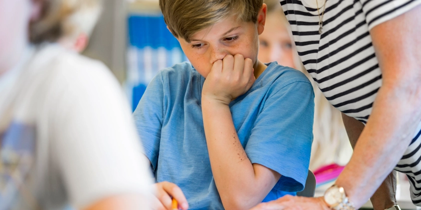En ung pojke sitter vid en skolbänk med en kvinna bakom sig.