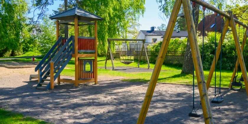 En lekpark med gungställningar och en lekställning med rutschkana,  omgiven av grönska.