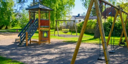 En lekpark med gungställningar och en lekställning med rutschkana,  omgiven av grönska.