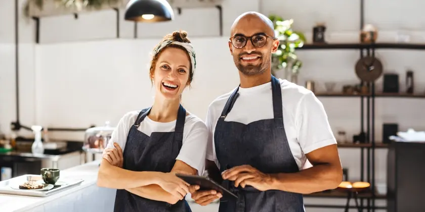 En kvinna och en man, båda med förkläden står i en restaurang och ser glada ut