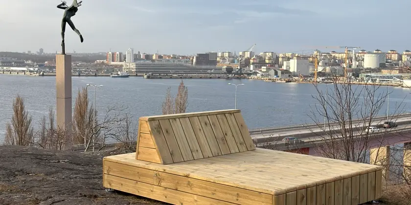 Solsäng i ljust trä står på en gräsplätt på Torsviksberget med utsikt över Stockholm och med en Millesstaty i bakgrunden