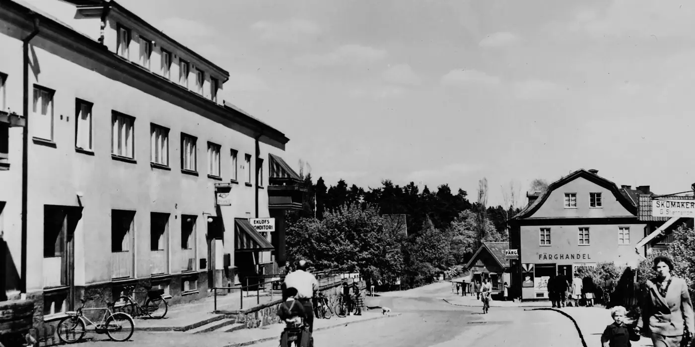 En man cyklar ner för Stockholmsvägen, förbi ett tvåvåningshus som är till vänster i bild. En kvinna går med ett barn till höger i bild. Bilden är svartvit från 40-talet