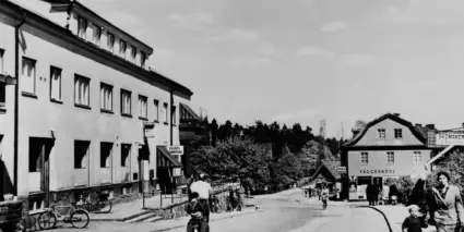 En man cyklar ner för Stockholmsvägen, förbi ett tvåvåningshus som är till vänster i bild. En kvinna går med ett barn till höger i bild. Bilden är svartvit från 40-talet
