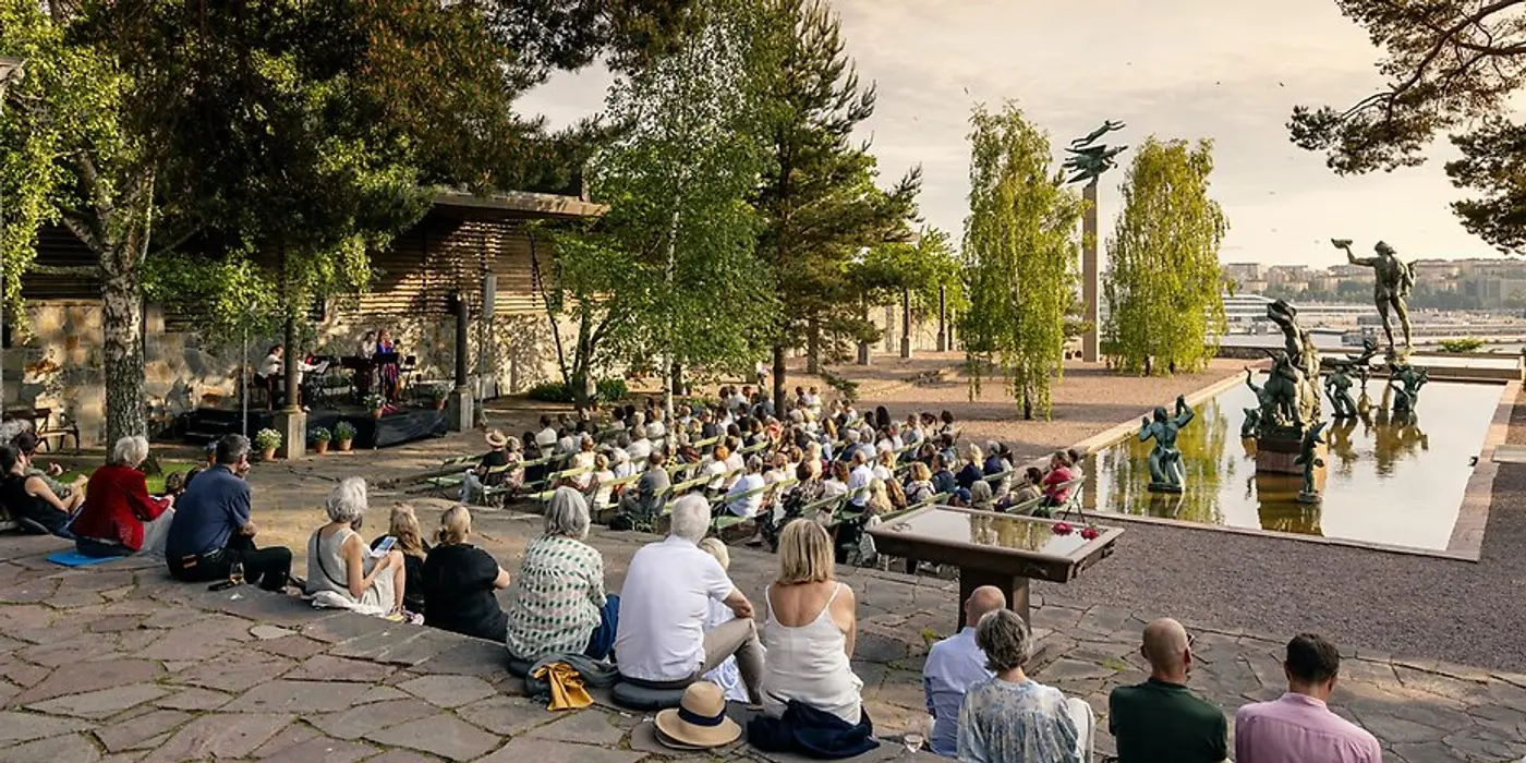 En grupp människor sitter vid en fontän på Millesgården och lyssnar på musiker som spelar på en scen till vänster i bilden. I omgivningen finns träd som ramar in platsen, i bakgrunden syns Milles statyer.