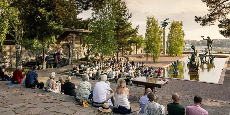 En grupp människor sitter vid en fontän på Millesgården och lyssnar på musiker som spelar på en scen till vänster i bilden. I omgivningen finns träd som ramar in platsen, i bakgrunden syns Milles statyer.