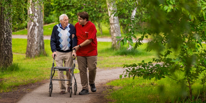 En man med rullator på promenad får hjälp av en manlig vårdpersonal.