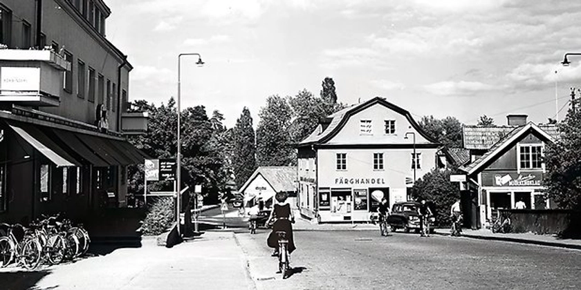 Ett svartvitt foto av en person som cyklar längs en gata omgiven av butiker.
