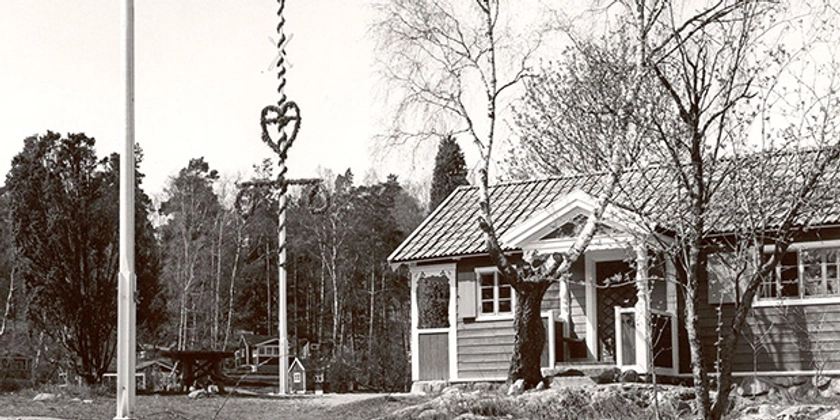 Ett svartvitt foto av en stuga med en flaggstång och en midsommarstång.