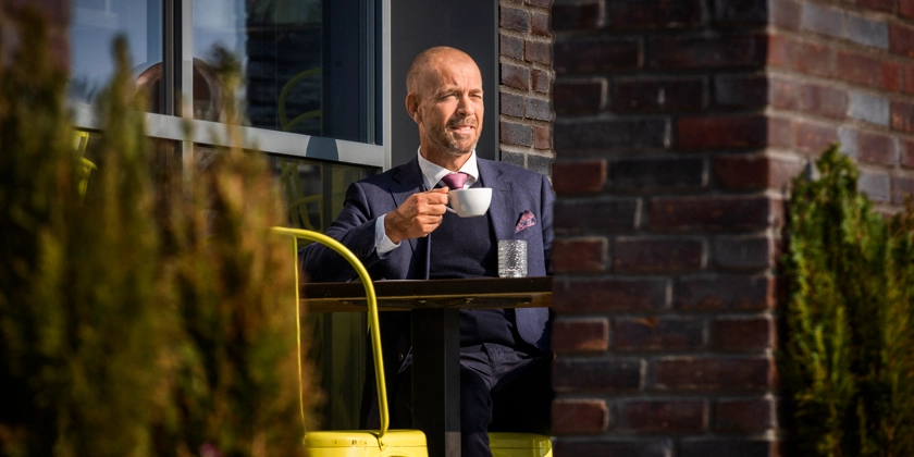 En man sitter i solen på en uteservering och dricker en kopp kaffe.