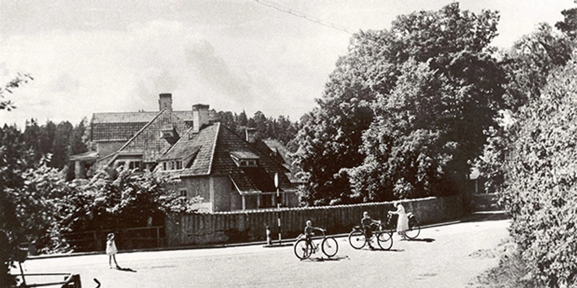 Ett svartvitt foto av människor med cyklar på en gata, framför ett stort hus.