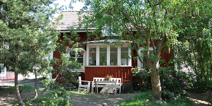 Ett litet rött hus i lummig trädgård med vita trädgårdsmöbler. 