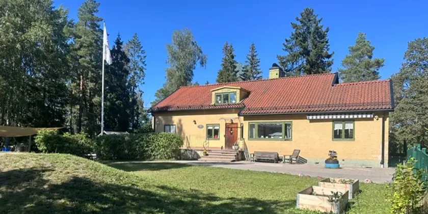 Gul villa med rött tak omgiven av stor gräsmatta och höga träd.