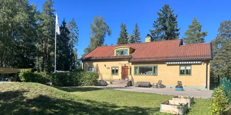 Gul villa med rött tak omgiven av stor gräsmatta och höga träd.