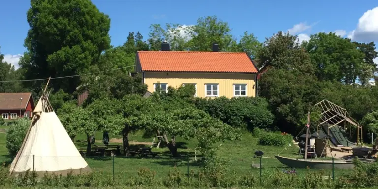 Ett tipi som står mitt på en äng, i bakgrunden en gul villa.
