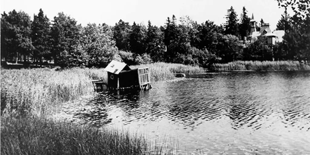 Ett svartvitt foto av en sjö med sjöbod och en stor villa i bakgrunden.