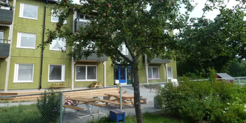 I förgrunden ett träd framför en uteplats vid ett trevåningshus.