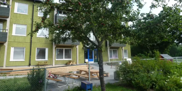 I förgrunden ett träd framför en uteplats vid ett trevåningshus.
