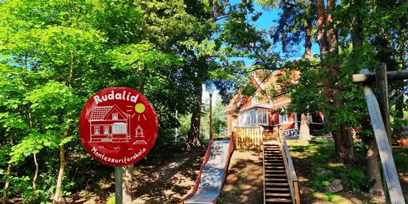 Röd villa skymtar fram bakom lummiga träd och rutschkana. 
