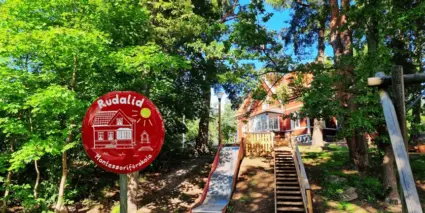 Röd villa skymtar fram bakom lummiga träd och rutschkana. 