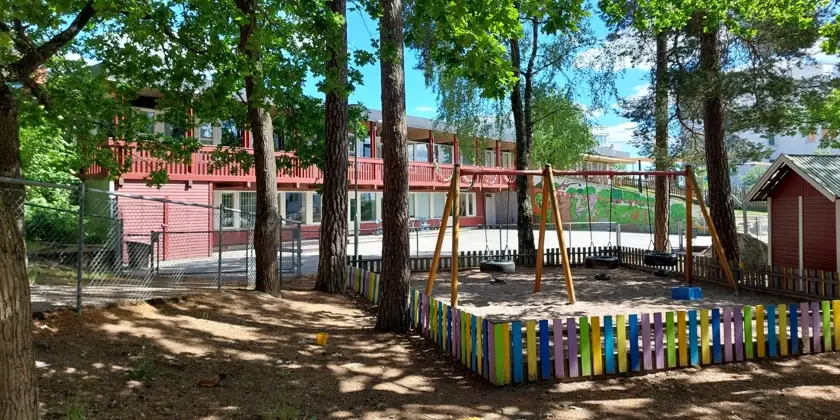 En park med en gunga och en två våningar hög förskolebyggnad i bakgrunden.