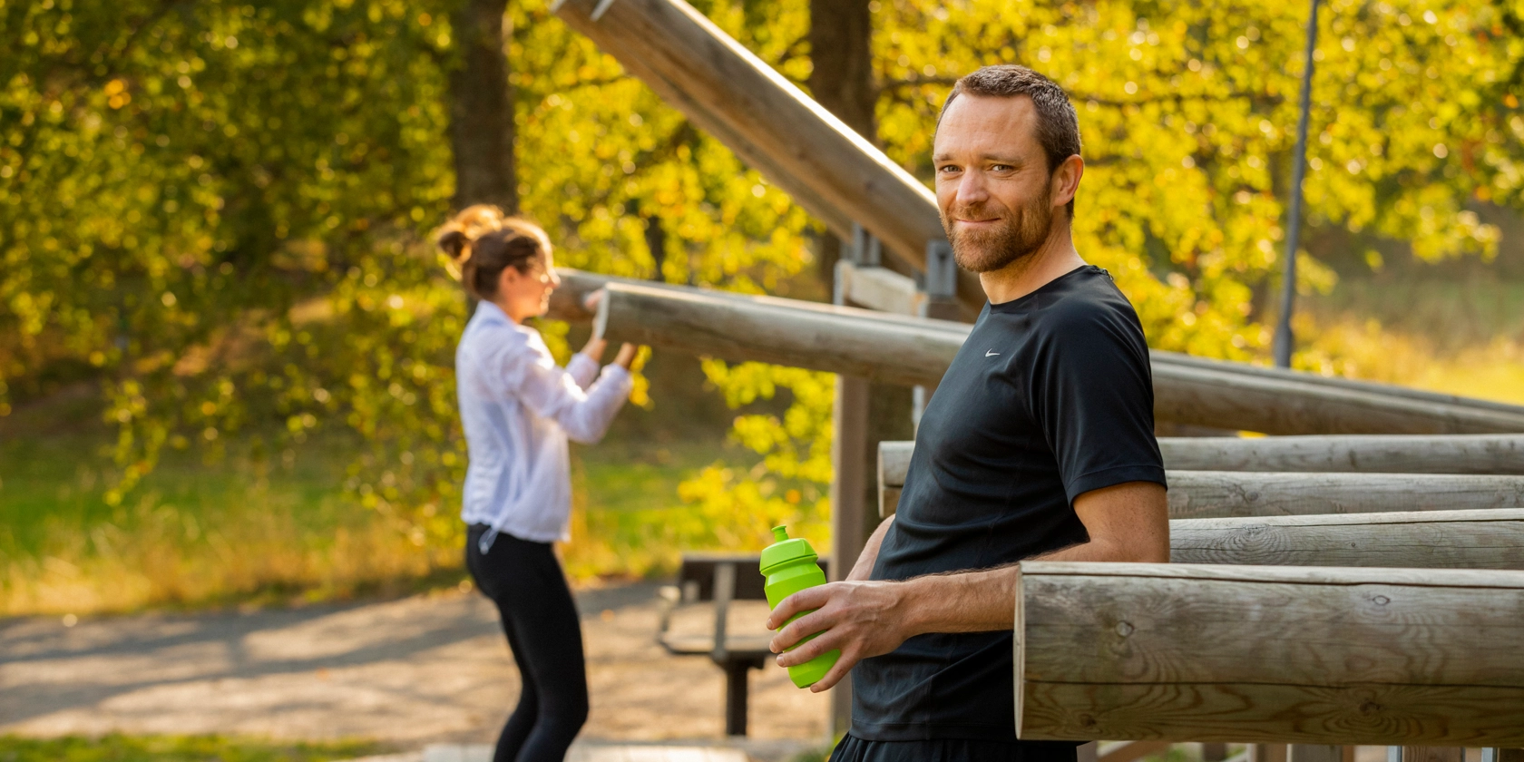 En kvinna och en man tränar på ett utegym, mannen tar en vätskepaus och ler mot kameran.